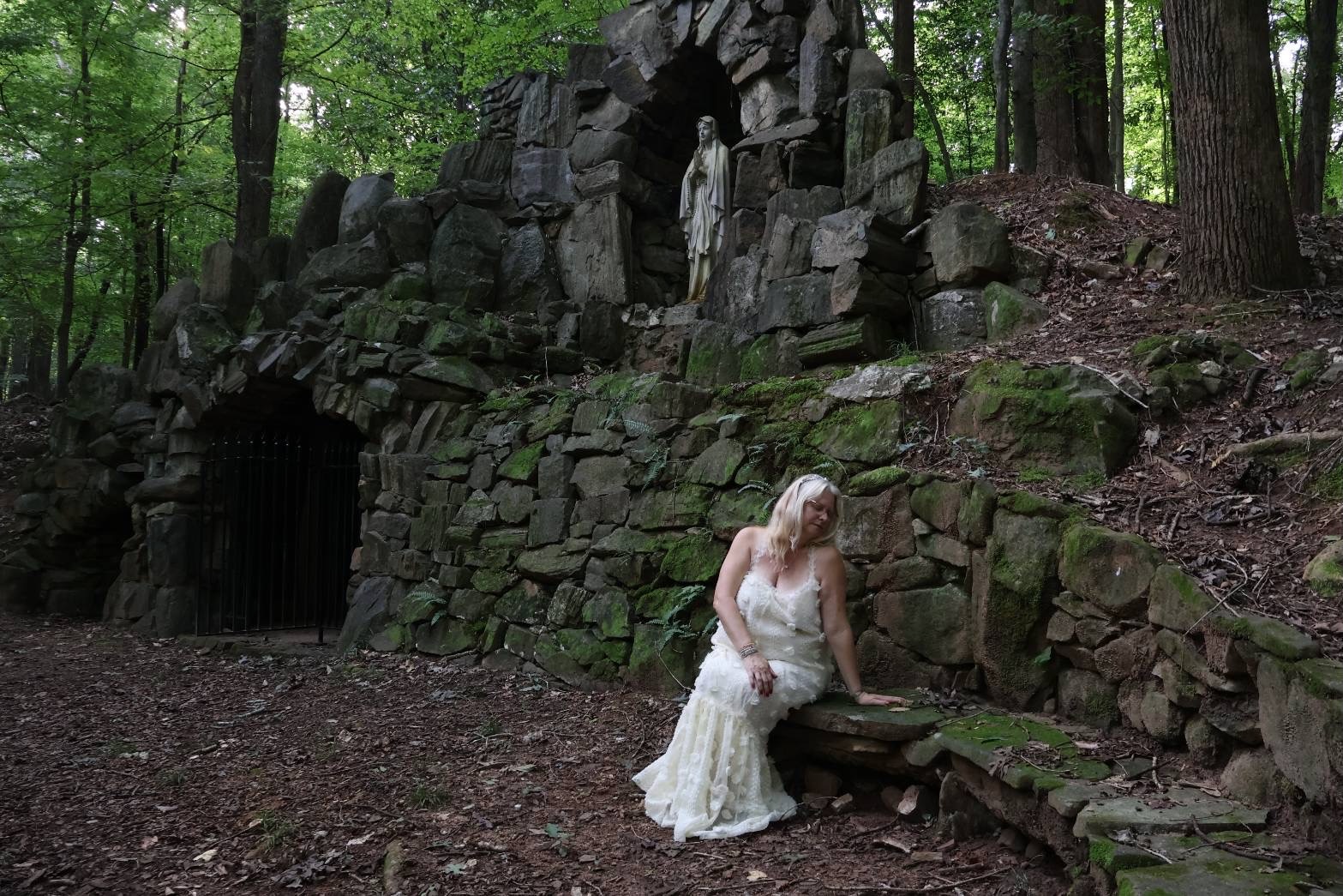 Beth Stuerman seated outdoors beside a stone wall in a wooded garden